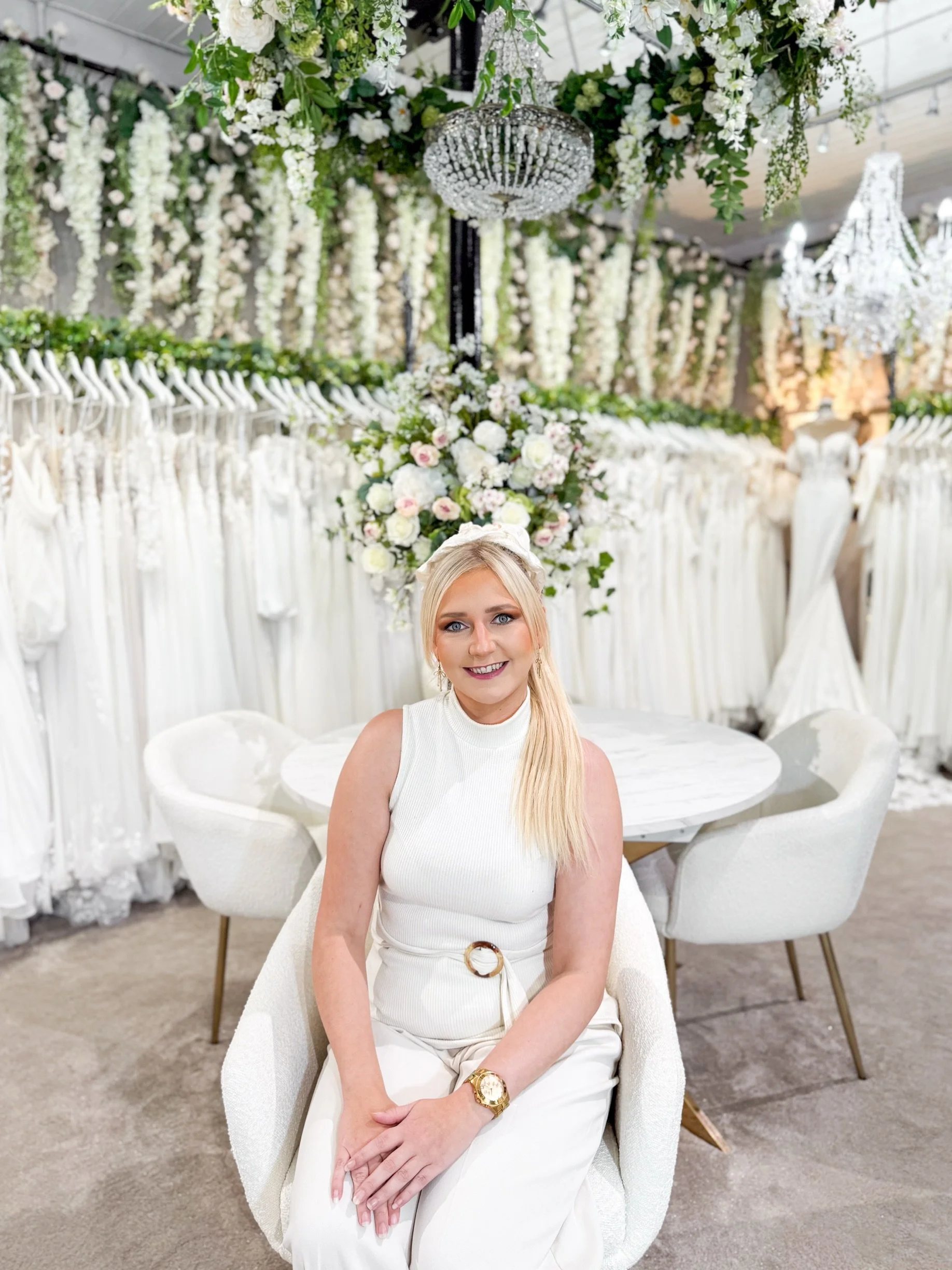 A woman with blonde hair wearing a navy blue sleeveless top and matching navy blue pants, standing in front of a floral arrangement and a mirror, smiling at the camera.