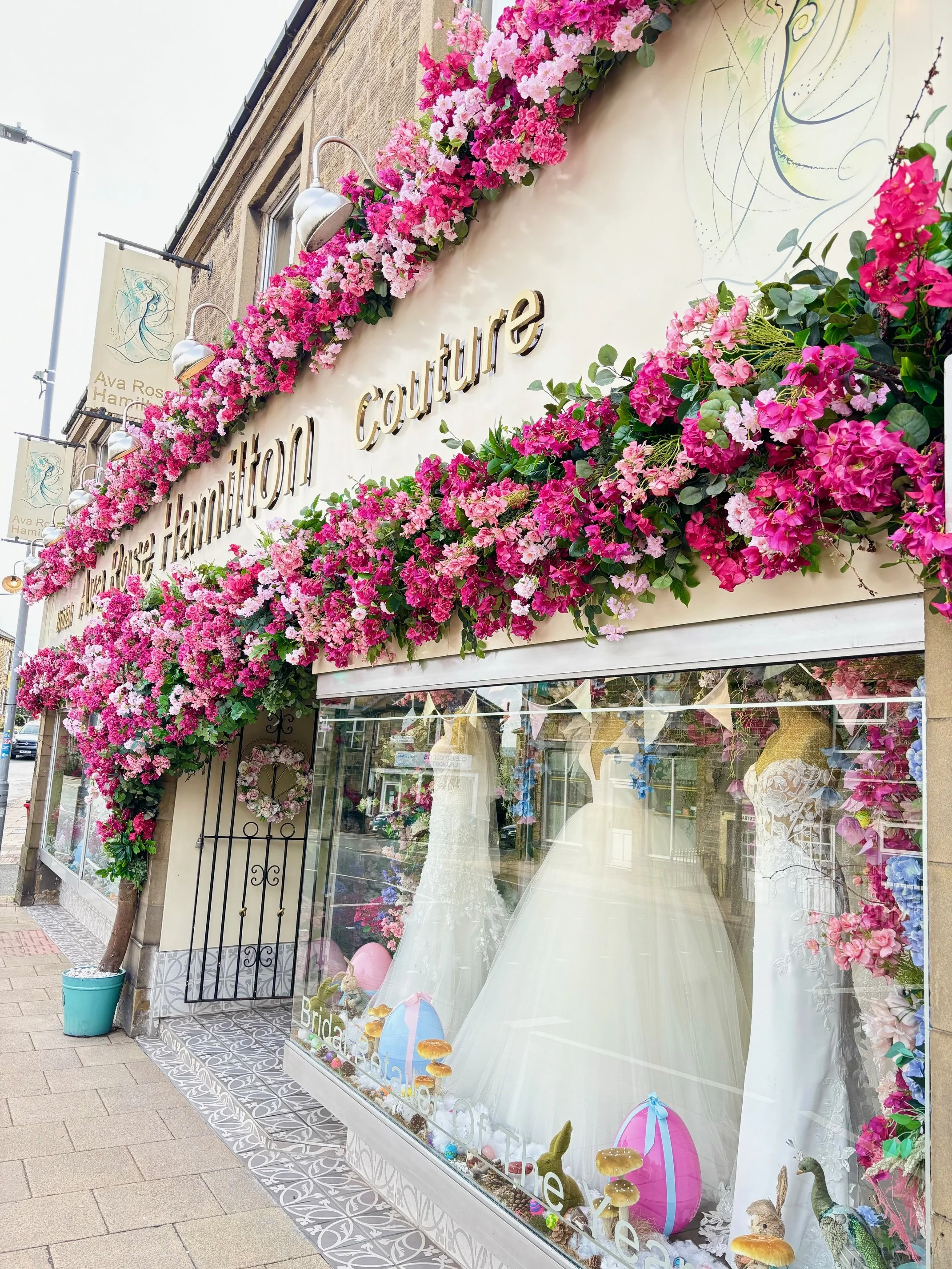 storefront decorated with pink and purple flowers, displaying wedding dresses inside a boutique named "Viva Rose Hamilton Couture" with Easter eggs and bunny decor in the window.