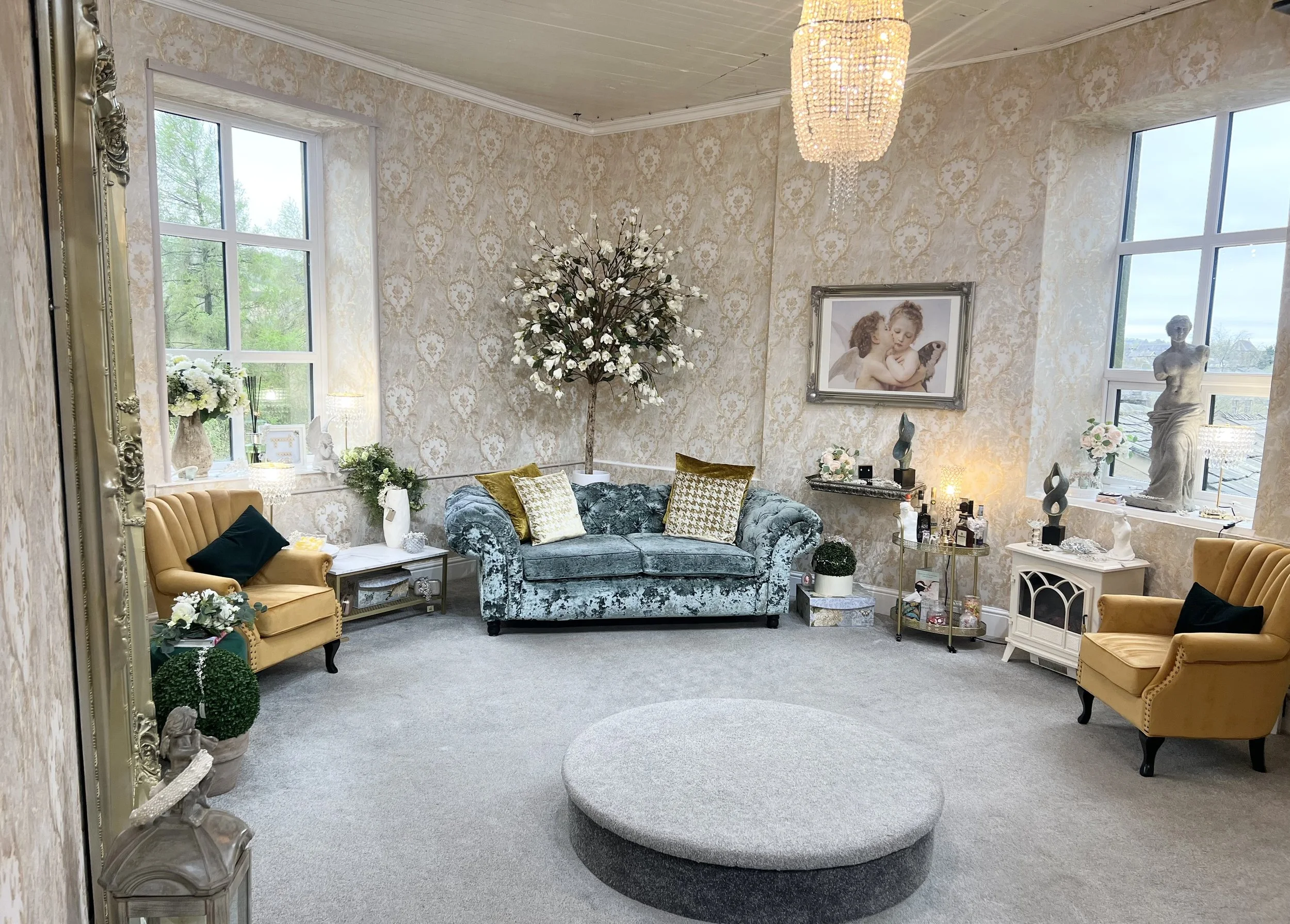 A living room with vintage decor, including a velvet sofa, yellow armchairs, floral wallpaper, and decorative statues. Large windows provide natural light, and the room has a chandelier.