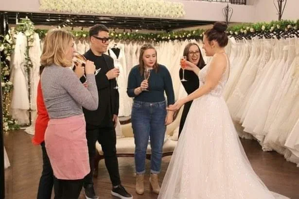 A bride wearing a white wedding gown holding hands with a woman in a black dress, surrounded by four women in casual clothing in a bridal shop decorated with wedding dresses and greenery.