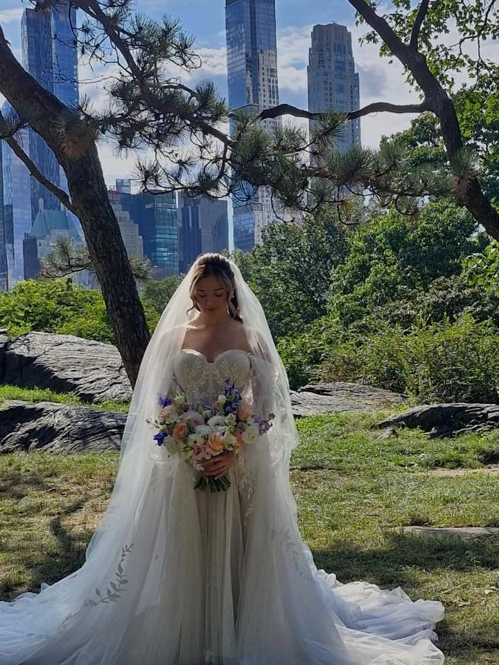 A bride in a wedding dress holding a bouquet of flowers, standing outdoors under trees with a city skyline in the background.