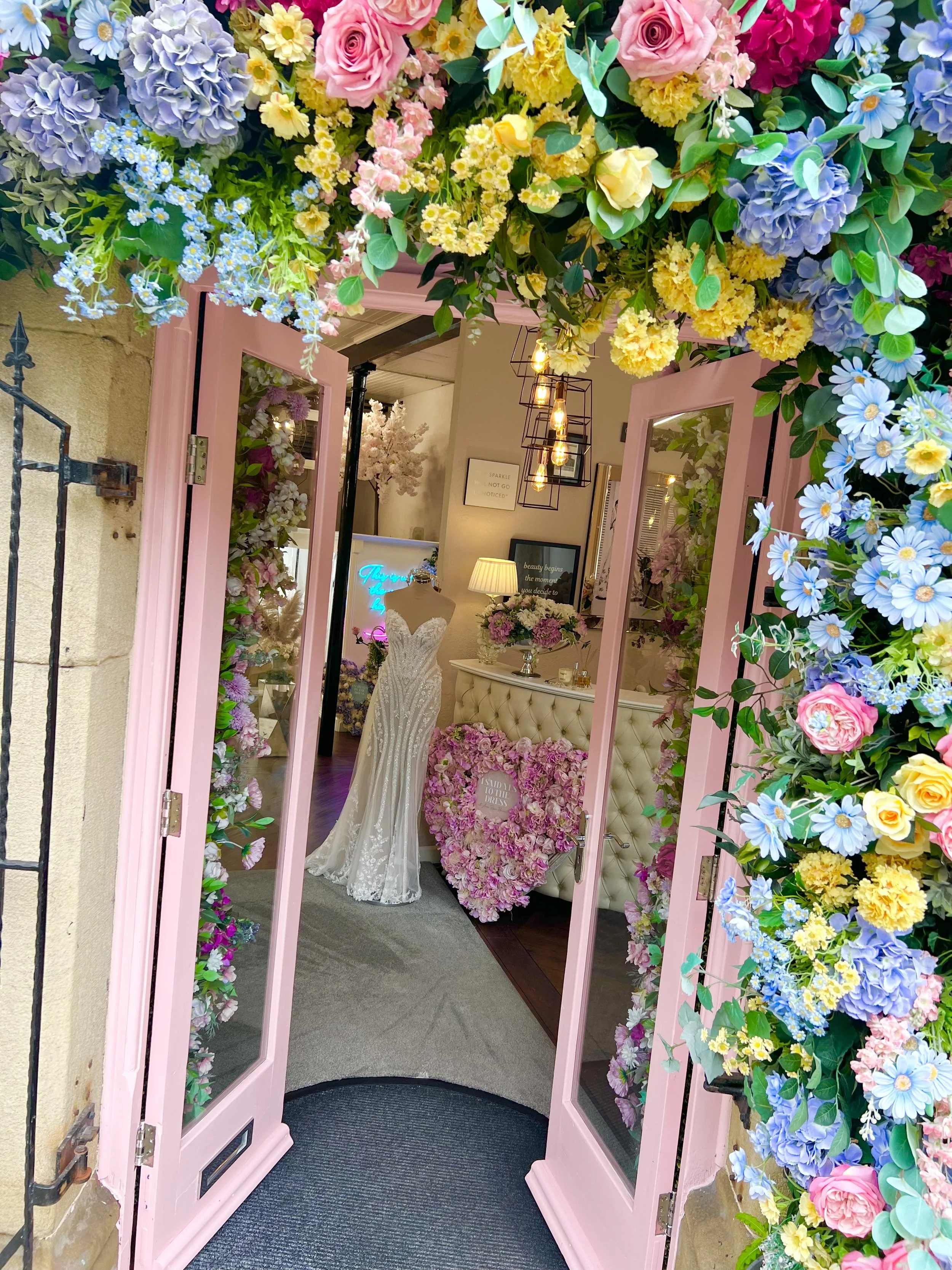 Pink floral decorated entrance to a boutique wedding dress shop, with a wedding gown displayed inside, surrounded by flowers and soft lighting.