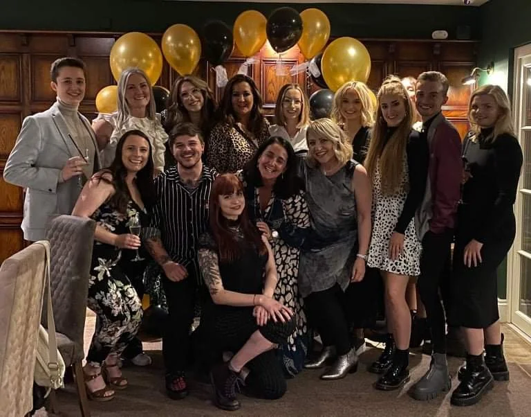Group of people celebrating at a party with black and gold balloons in the background.