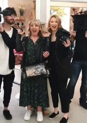 Four people in a kitchen, smiling and posing for a photo. Two women in the center are excited, one holding a black object, the other with a purse. Two men are partially visible on either side, one on a virtual reality headset.