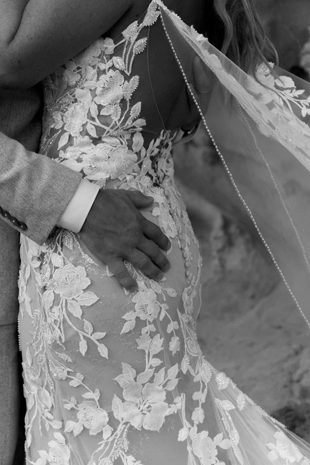Close-up of a bride and groom, with the groom gently holding the bride's pregnant belly. The bride is wearing a lace wedding dress and a veil.