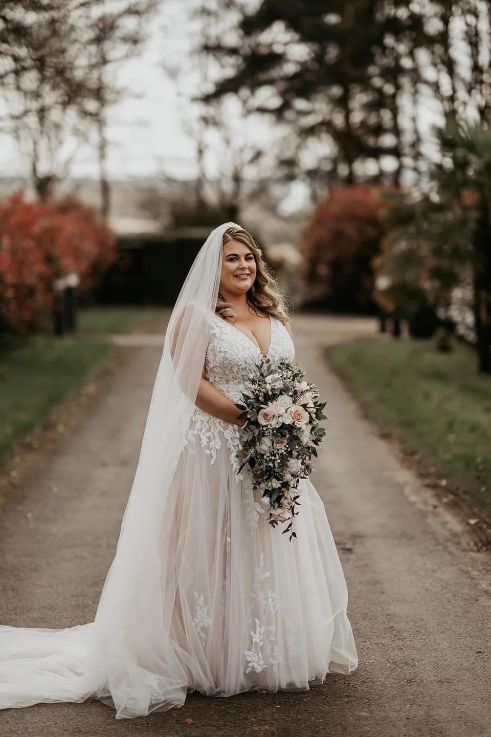 A bride in a white wedding gown with lace details and a long train, holding a cascading bouquet of pink and white roses with greenery, standing on a pathway in a park with autumn-colored trees.
