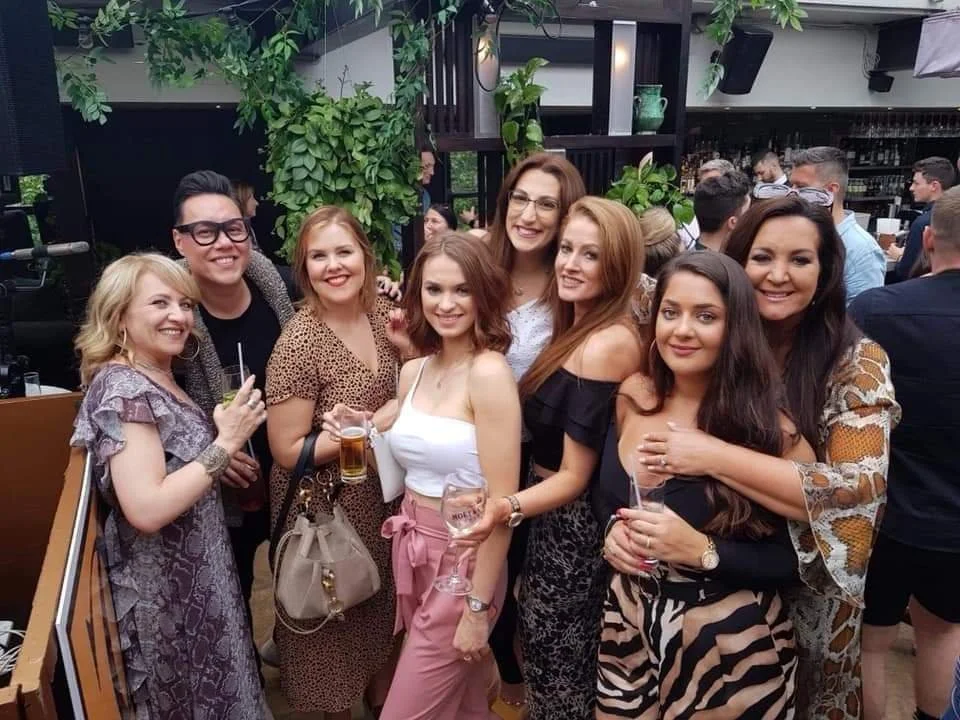 Group of women and one man at a social gathering, posing for a photo, some holding drinks, in an indoor venue with greenery and other people in the background.