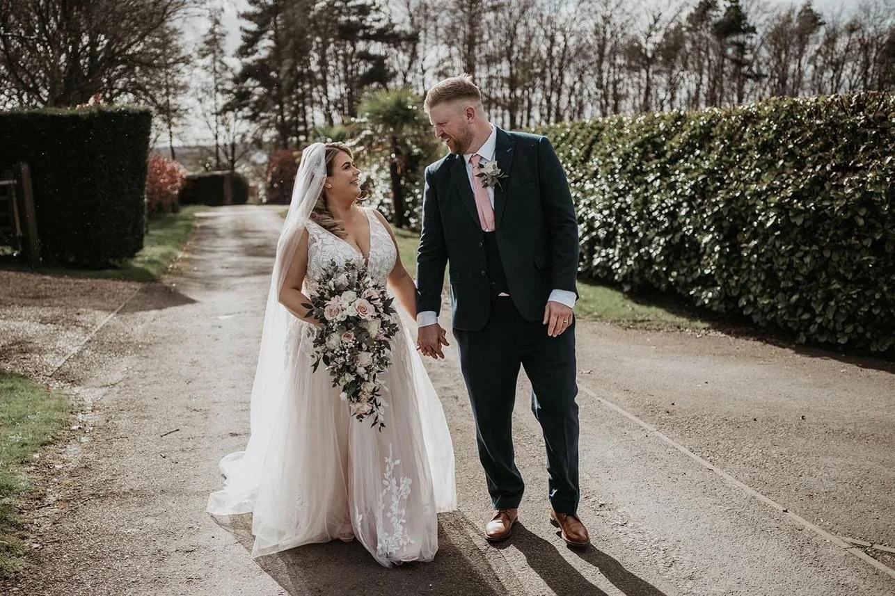 A bride and groom walk hand in hand outdoors on a paved path, smiling at each other. The bride wears a white wedding gown with lace details and a veil, holding a bouquet. The groom wears a dark suit with a pink tie and a boutonniere. There are bushes