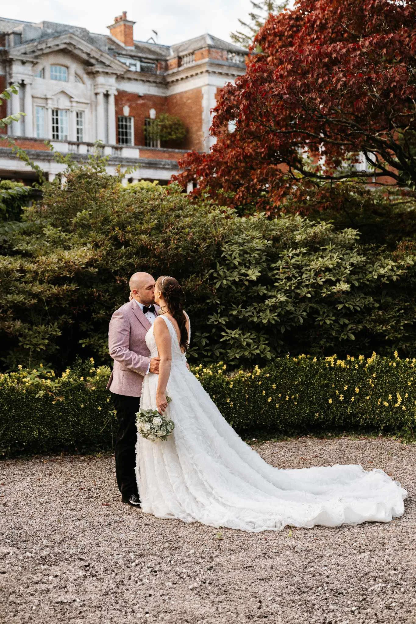 A newlywed couple sharing a kiss outdoors on their wedding day, with lush green foliage and a large red tree in the background, and a historic mansion or estate building behind them.