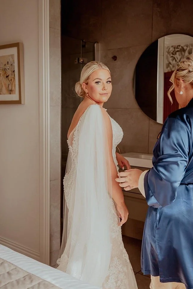 Bride in a wedding gown looks over her shoulder as another woman, possibly her maid of honor or a stylist, adjusts her dress in a bathroom or bedroom.