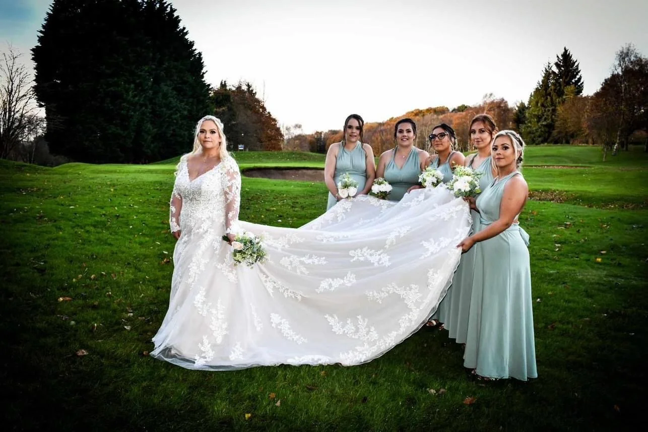 Bride in a white lace wedding gown holding a bouquet, standing outdoors on a grassy field with six bridesmaids in light blue dresses holding bouquets, posing for a photo during a wedding celebration.