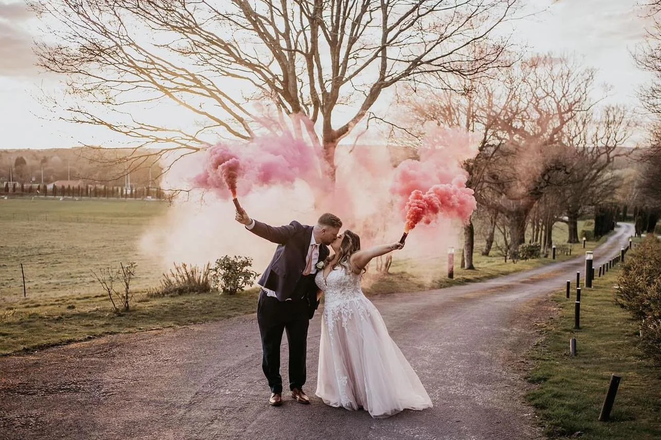 Newlywed couple embracing and kissing outdoors on a park path, holding pink smoke bombs, with a large leafless tree and open fields in the background during late afternoon or early evening.