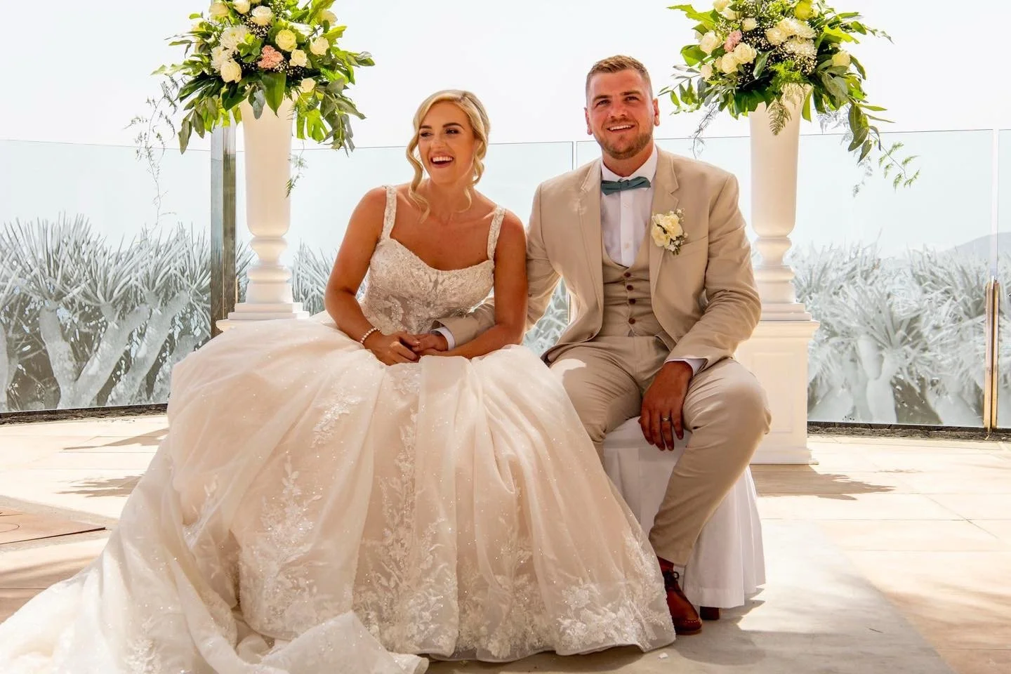 A bride and groom sitting together on a white bench during their wedding ceremony. The bride is wearing a white lace wedding gown, and the groom is in a beige suit with a bow tie. They are smiling and holding hands, with large floral arrangements pos