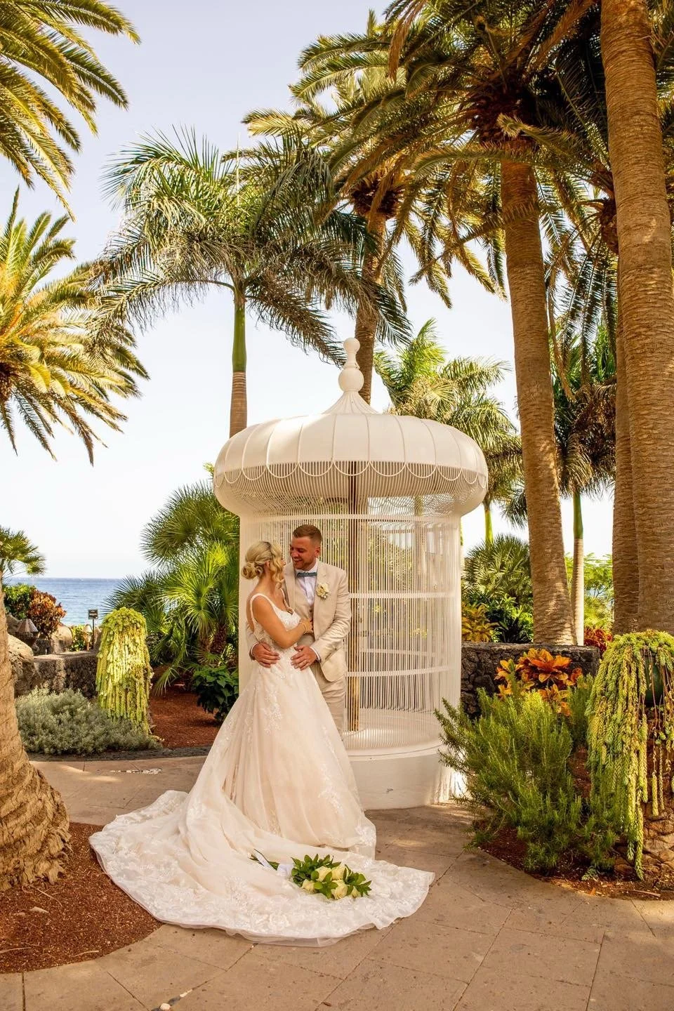 A bride and groom standing in front of a white gazebo surrounded by palm trees and tropical plants, with the ocean visible in the background, during a wedding ceremony.