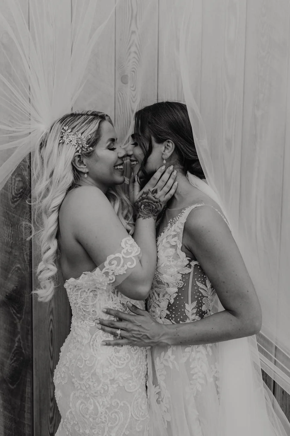 Two women in wedding dresses sharing an intimate moment, touching noses and smiling, with one holding the other's face, in black and white.