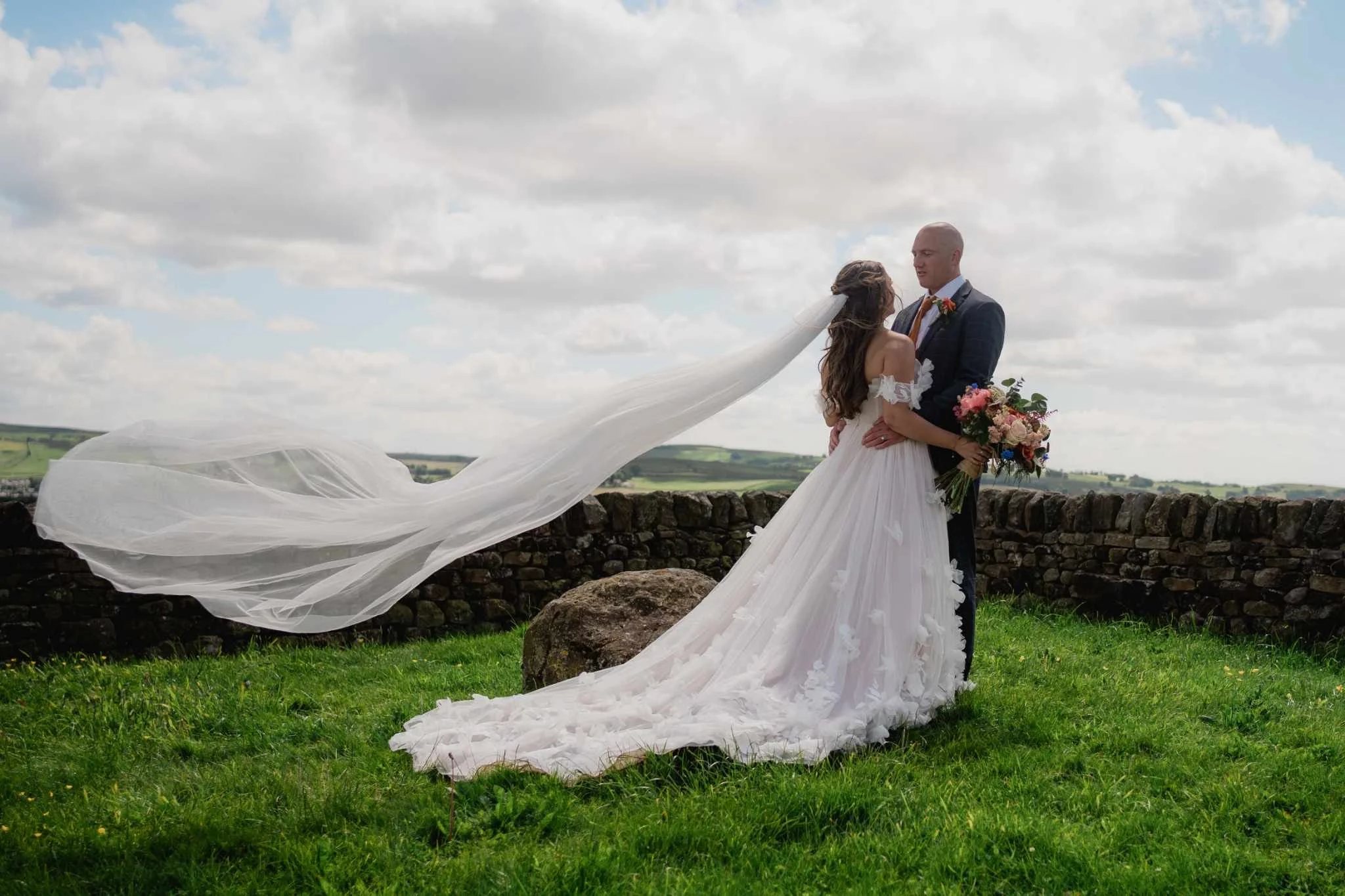 A bride and groom standing on grass in front of a stone wall, embracing and looking at each other during a wedding photoshoot outdoors. The bride is holding a bouquet of colorful flowers and has a long flowing veil in the wind.
