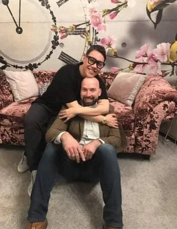 Two men smiling and sitting on a pink velvet sofa, with one standing behind and hugging the other. Behind them are pink flowers and a large clock with Roman numerals on the wall.