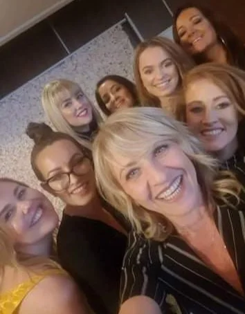 Group of eight women taking a selfie together indoors, smiling and looking happy.