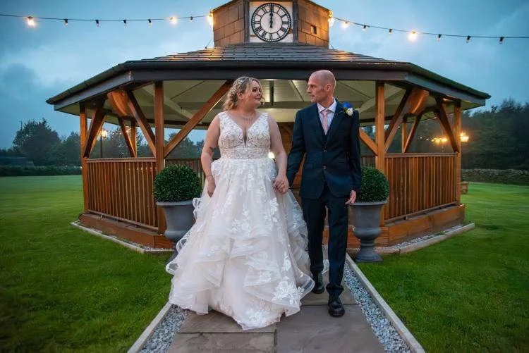 Bride and groom holding hands, walking towards the camera, in front of a wooden gazebo with a clock tower, during their wedding celebration at dusk.