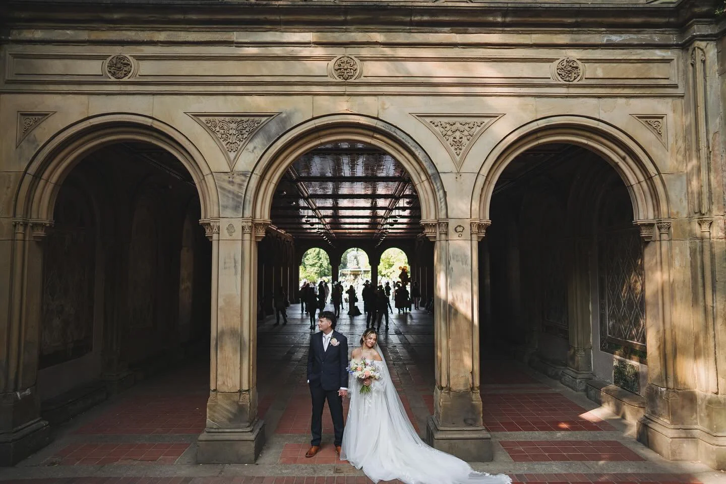 Bride and groom holding hands under a stone archway, with wedding guests in the background.