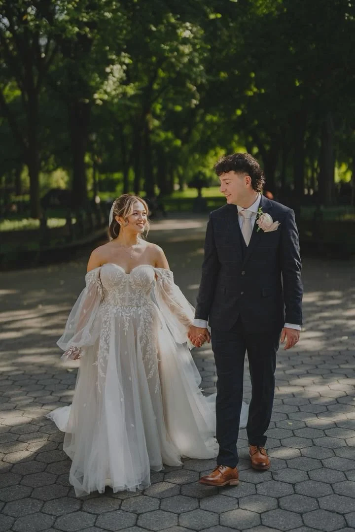 A bride and groom holding hands and walking outdoors on a sunny day, surrounded by green trees.