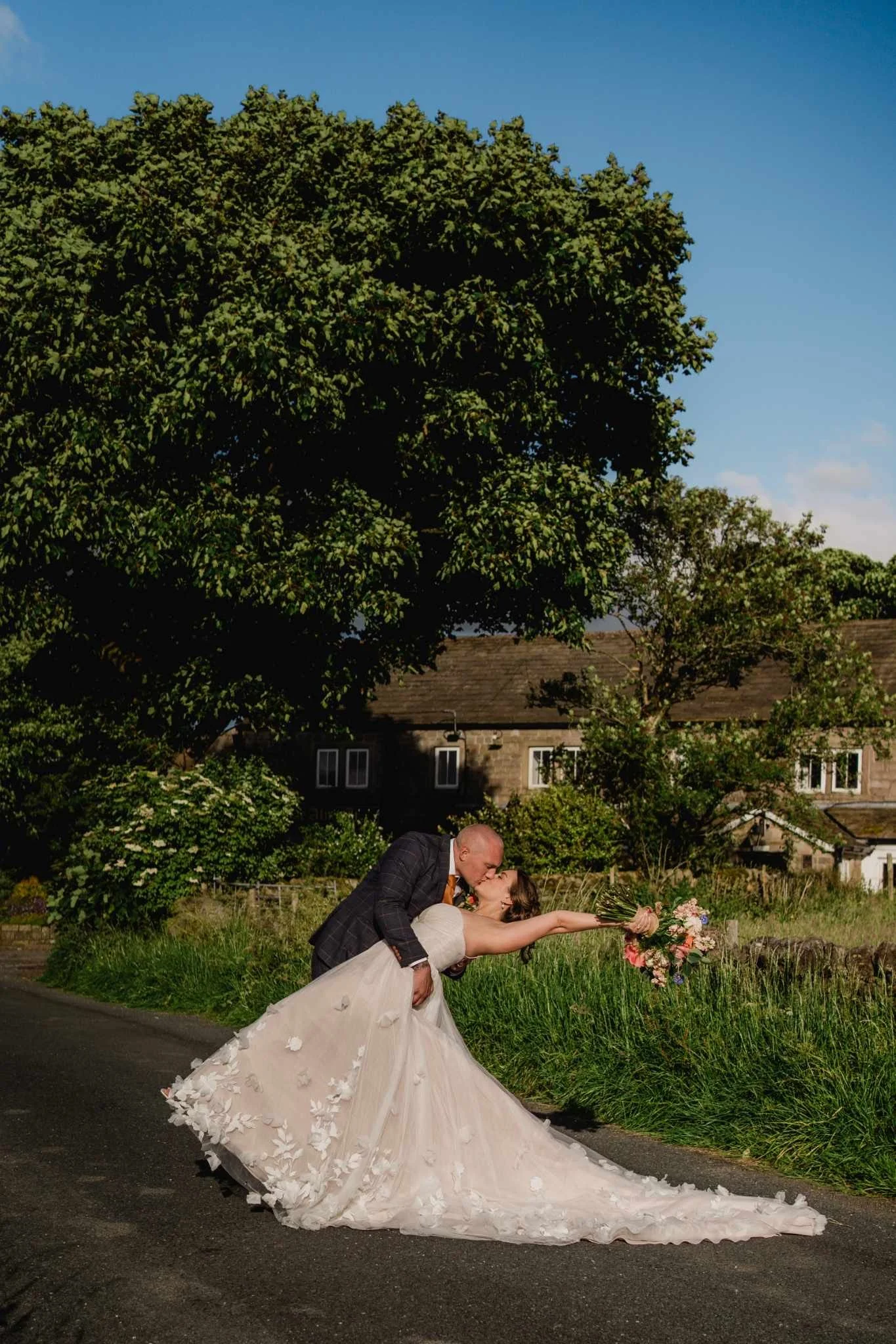 A newlywed couple sharing a kiss, with the groom dipping the bride while she holds a bouquet of pink and white flowers, standing on a rural road with greenery and trees in the background.