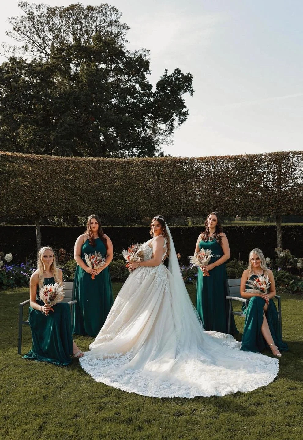 A bride in a white wedding gown and veil stands outdoors with four bridesmaids in teal dresses, holding bouquets, in a garden setting with trees and bushes in the background.