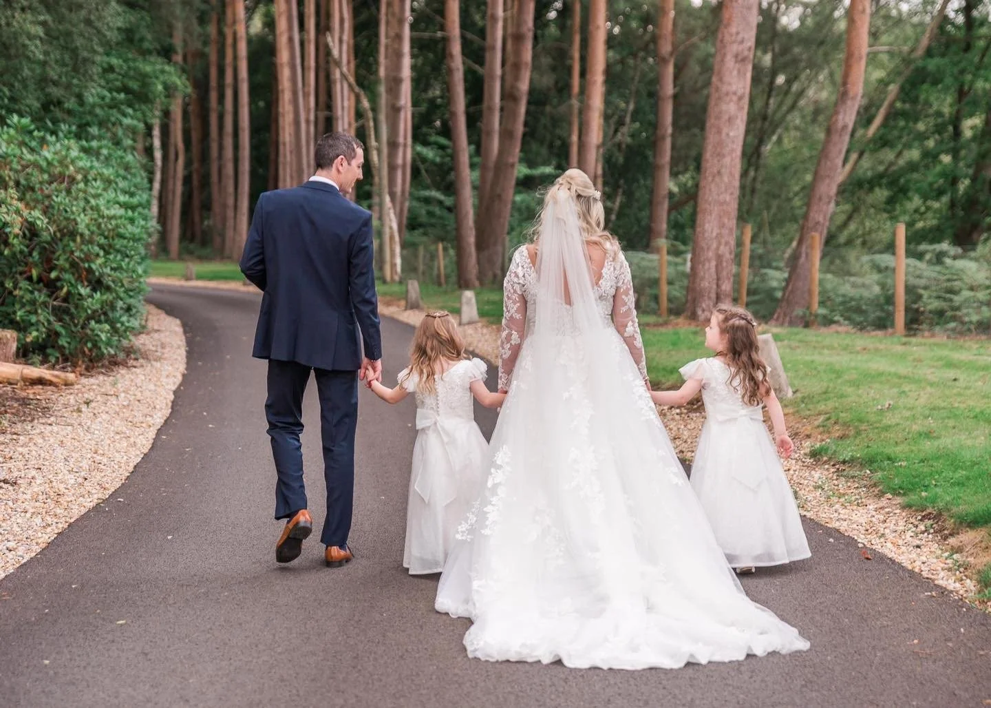 A bride and groom walking hand in hand with two young girls in white dresses on a paved path in a forested area.