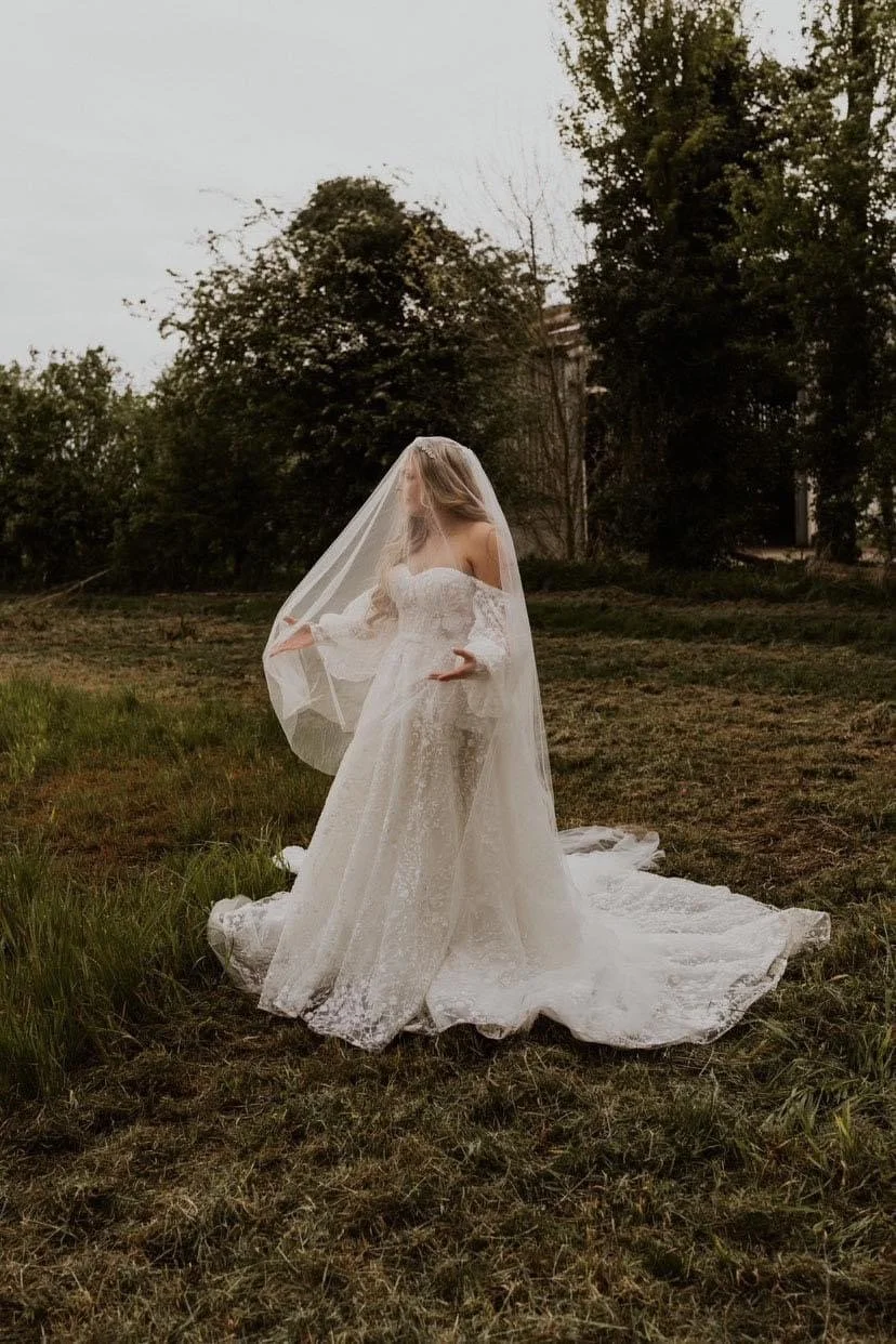 A bride in a white wedding dress and veil standing outdoors on grass with trees in the background.