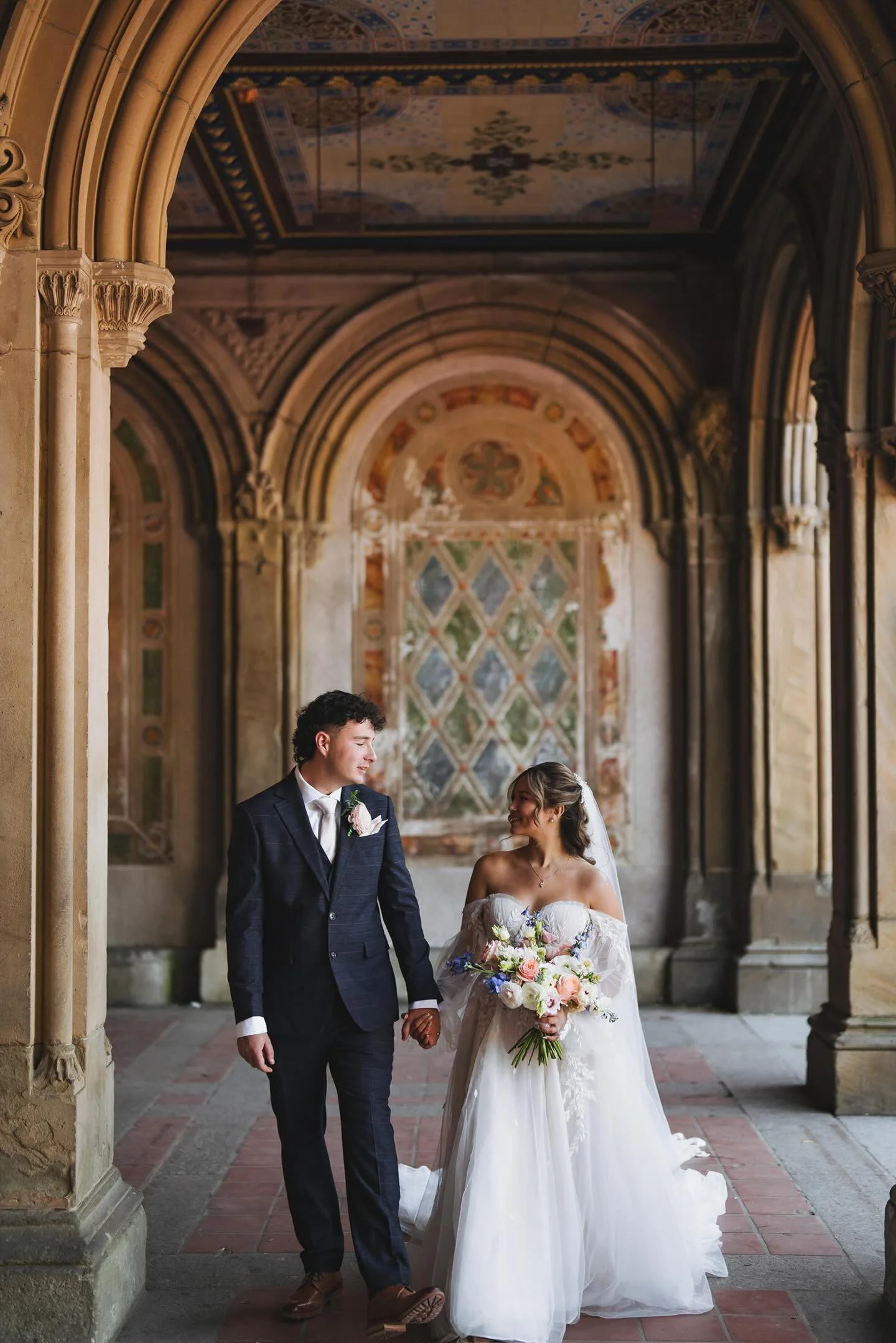 A bride and groom holding hands, walking through an ornate, arched, historic building with detailed frescoes and stained glass windows.