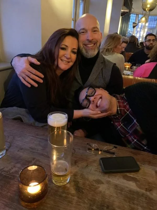 Three people smiling and hugging in a restaurant setting, with drinks and a candle on the table.