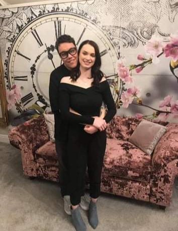Two women standing close together in front of a vintage floral wallpaper with a large clock design, in a decorated room with a floral velvet sofa and pink flowers.
