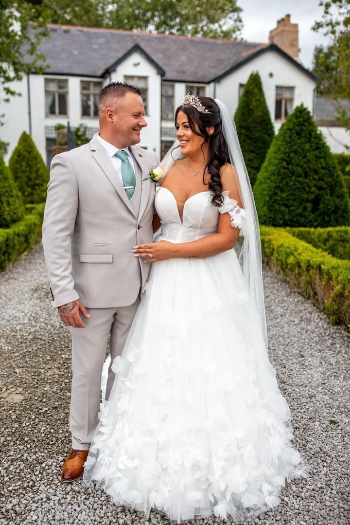 Bride and groom smiling at each other outdoors on their wedding day, with a white house in the background and trimmed garden bushes around them.