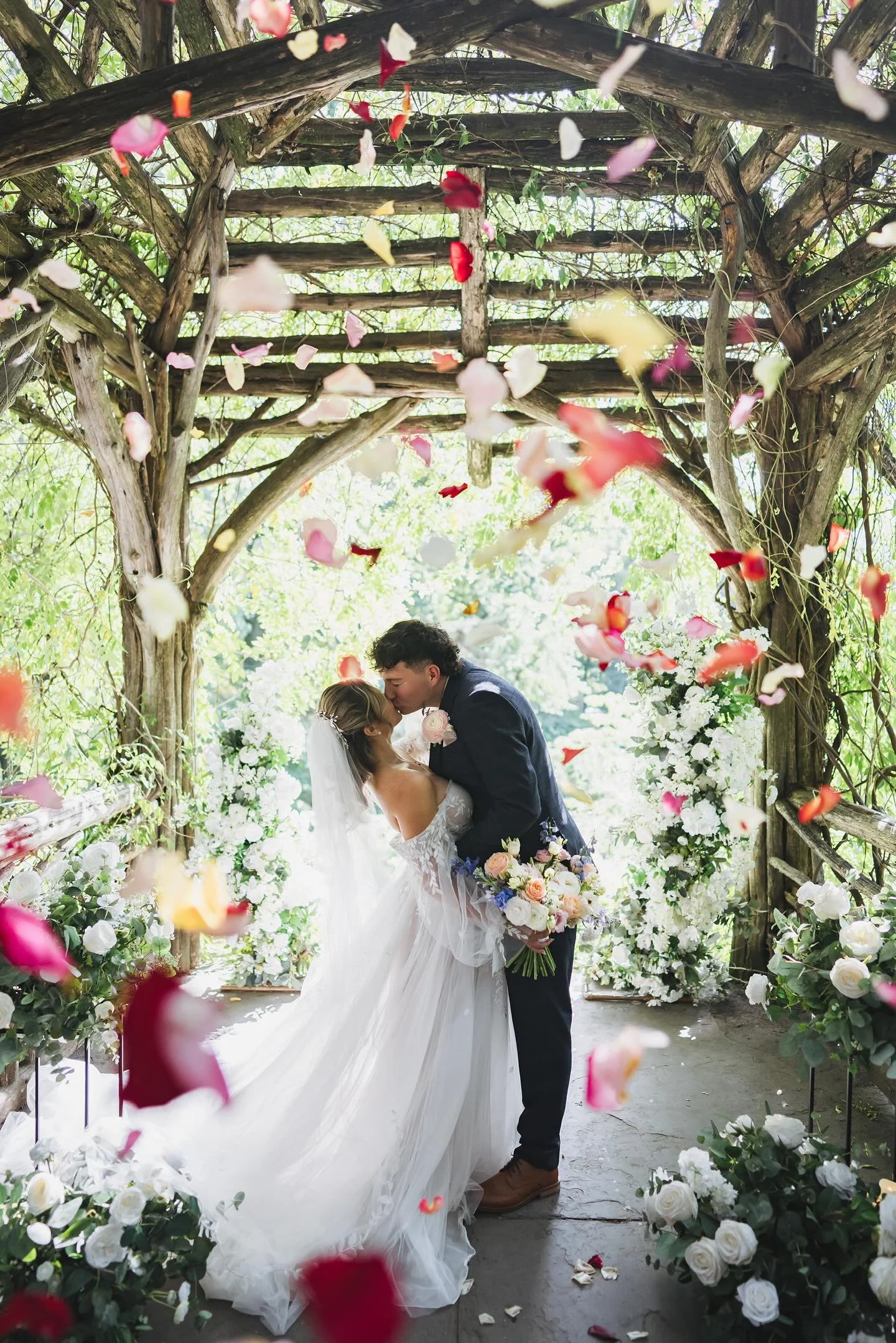 A bride and groom sharing a kiss at their wedding beneath a wooden arbor decorated with white flowers. Rose petals are falling around them, and the bride holds a bouquet of pastel and blue flowers. The setting is outdoor, lush with greenery.