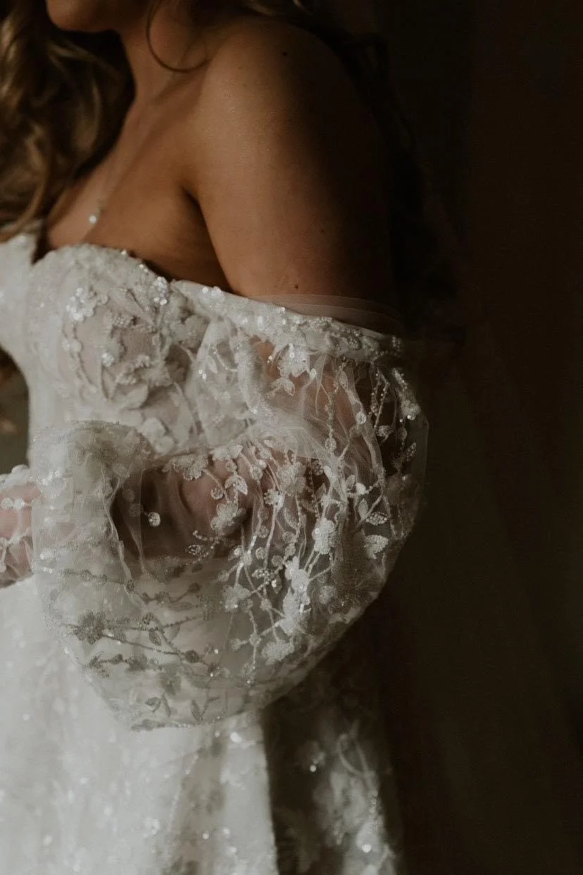 Close-up of a woman wearing a white lace wedding dress with floral embroidery, partially off-shoulder, with dark hair cascading down, in a dimly lit setting.