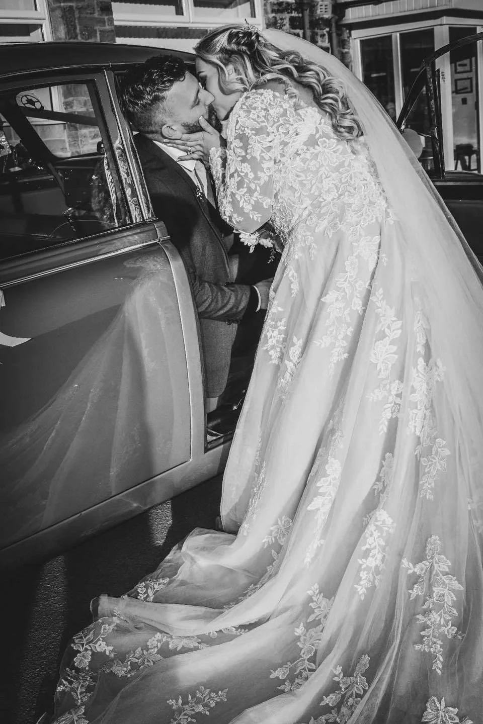 Black and white photo of a bride and groom sharing a kiss beside a vintage car, with the bride lifting her veil and wearing a lace wedding dress.