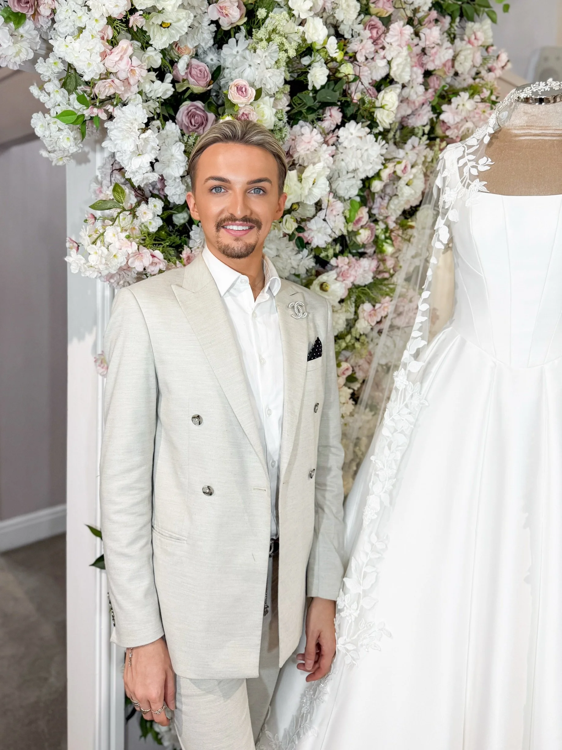 A person with blonde hair, blue eyes, and a goatee taking a selfie in a bridal shop, with elegant wedding dresses and floral decorations in the background.