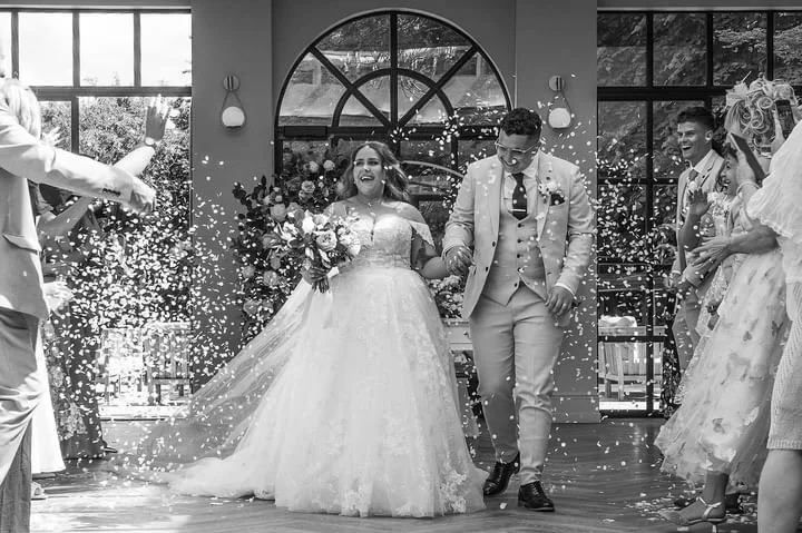 Bride and groom holding hands walking through a shower of confetti, surrounded by friends and family in a decorated venue with large windows.