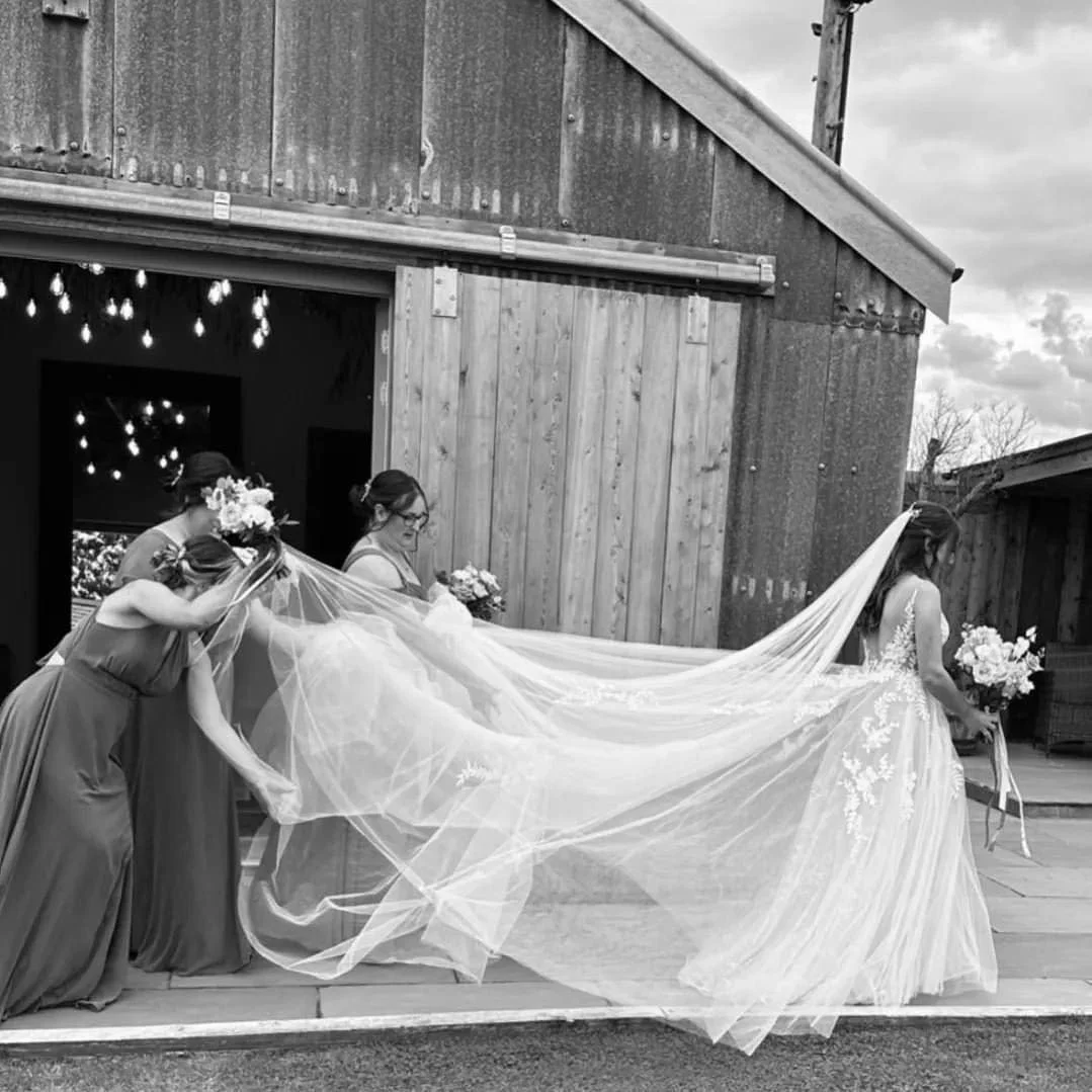 A bride in a wedding dress holding a bouquet and three bridesmaids in dresses holding the bride's veil outside a rustic building.