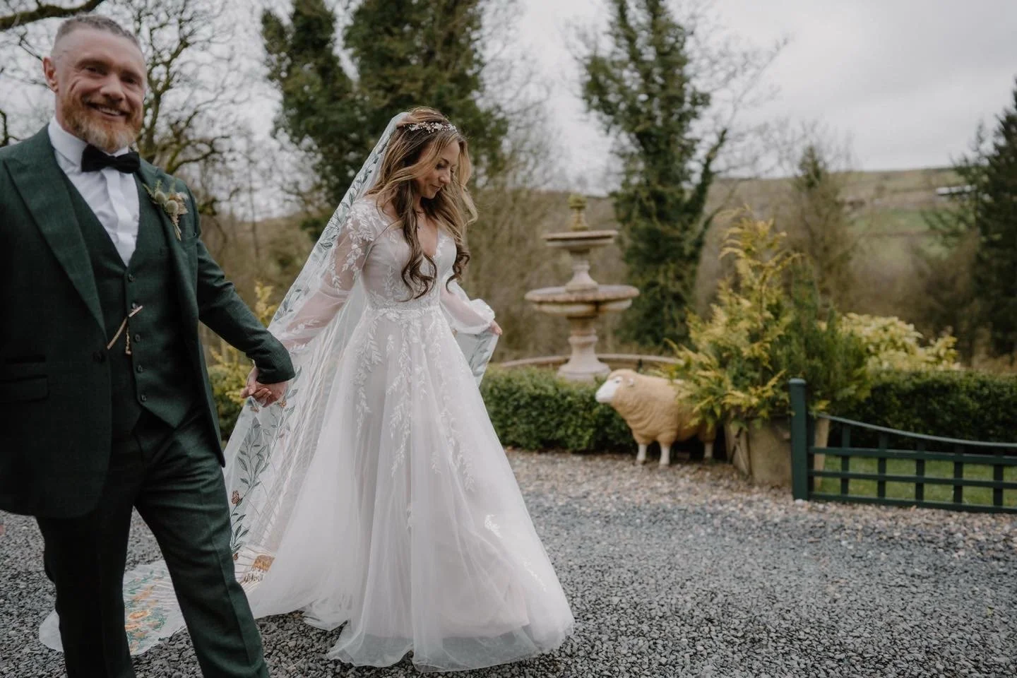 Bride and groom holding hands outdoors during wedding, the bride in a white gown with lace details and veil, the groom in a dark suit with a bow tie, trees and shrubbery in the background, overcast sky.