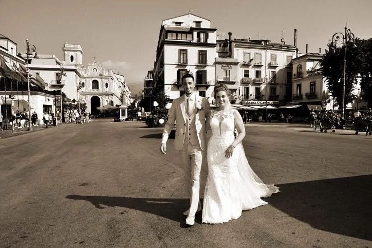 A newlywed couple in wedding attire walking on a wide city street in daytime, with historic buildings and storefronts in the background.