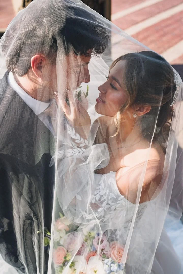 A bride and groom are close under a wedding veil, gazing into each other's eyes, with the bride holding a bouquet of pink and cream flowers.