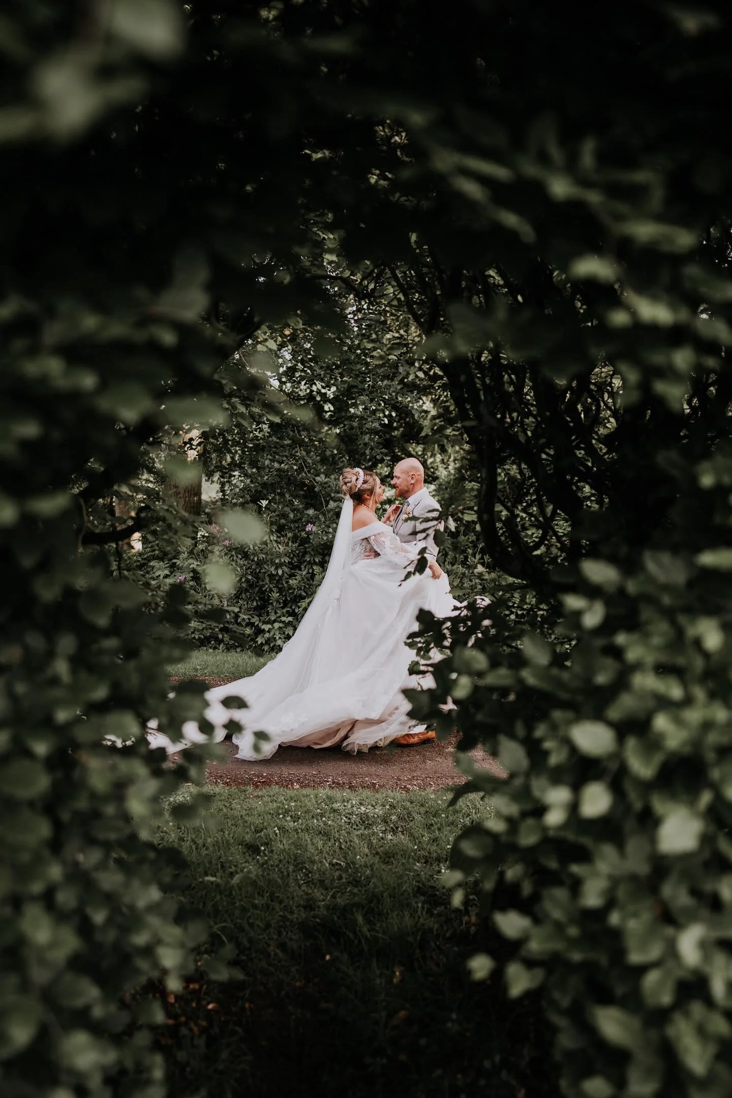 Bride and groom dancing outdoors, viewed through bushes, surrounded by greenery in a natural setting.