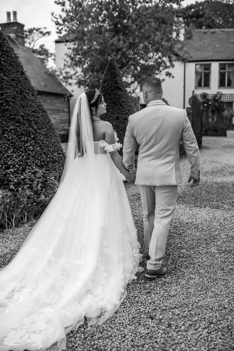 A bride and groom walking hand-in-hand outdoors on a gravel path, with the bride wearing a strapless wedding gown and veil, and the groom in a suit, surrounded by trees and houses.