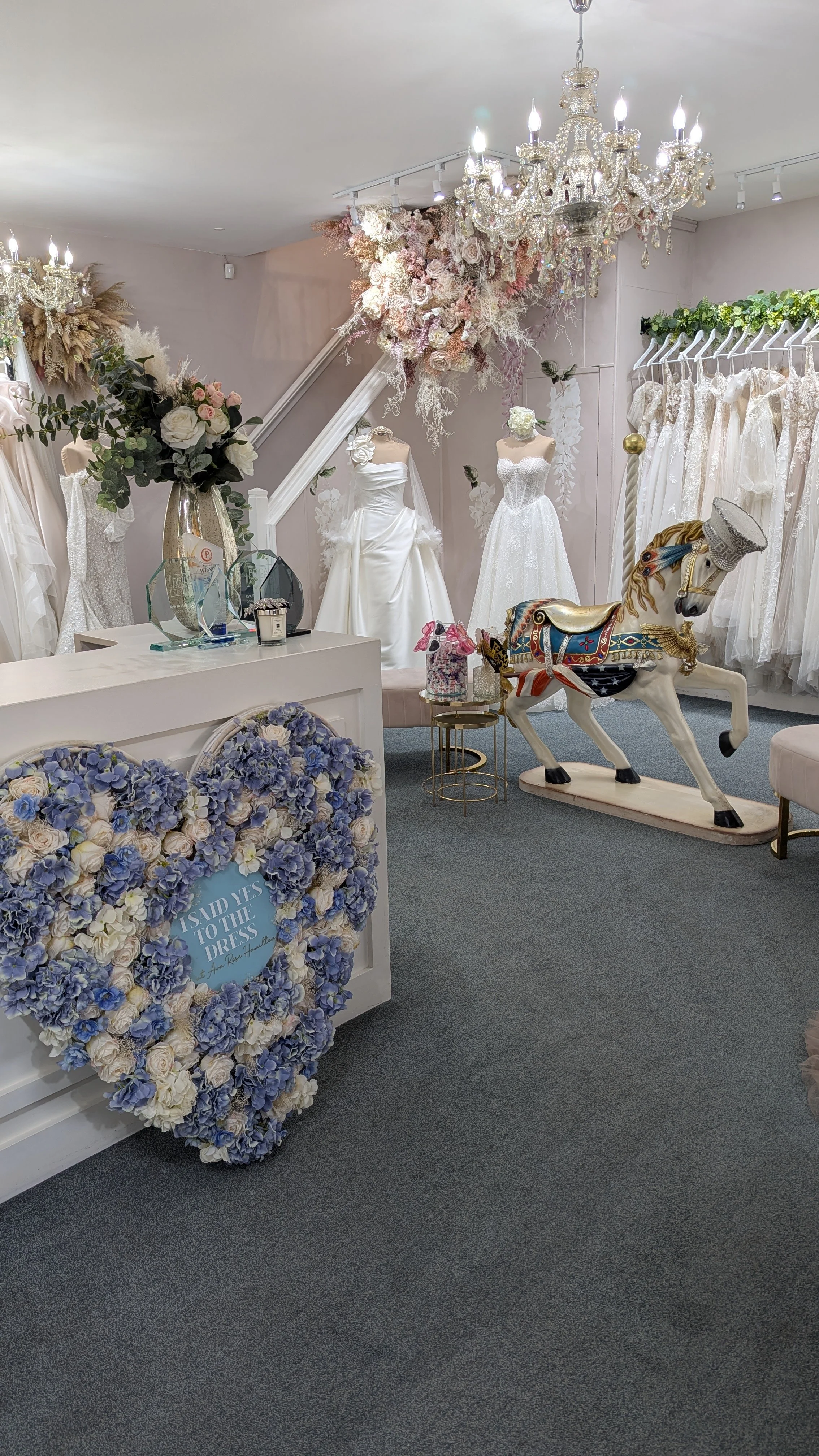 Interior of a bridal boutique with wedding dresses on mannequins, floral arrangements, a chandelier, and a vintage carousel horse.
