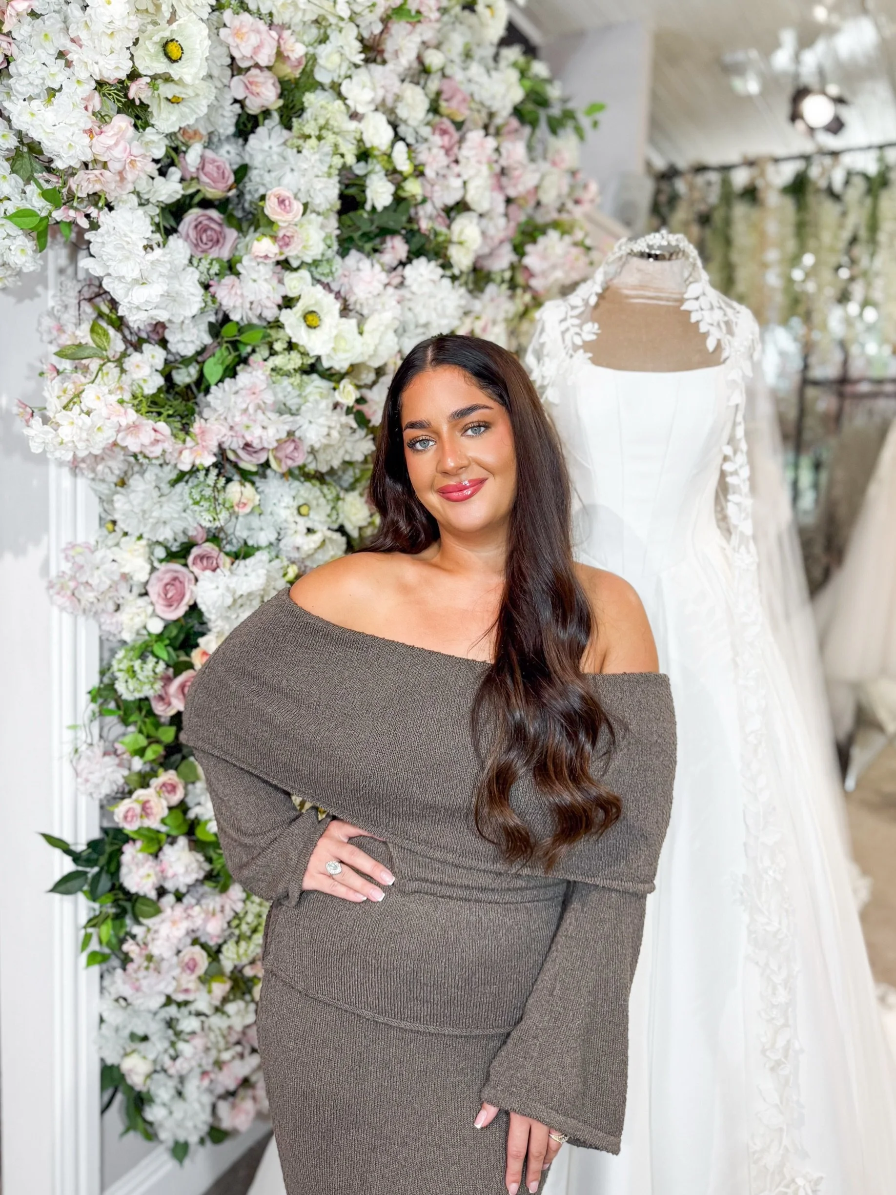 A woman with long dark hair, blue eyes, and light makeup, wearing a black sleeveless top, standing in front of a floral background and taking a selfie.