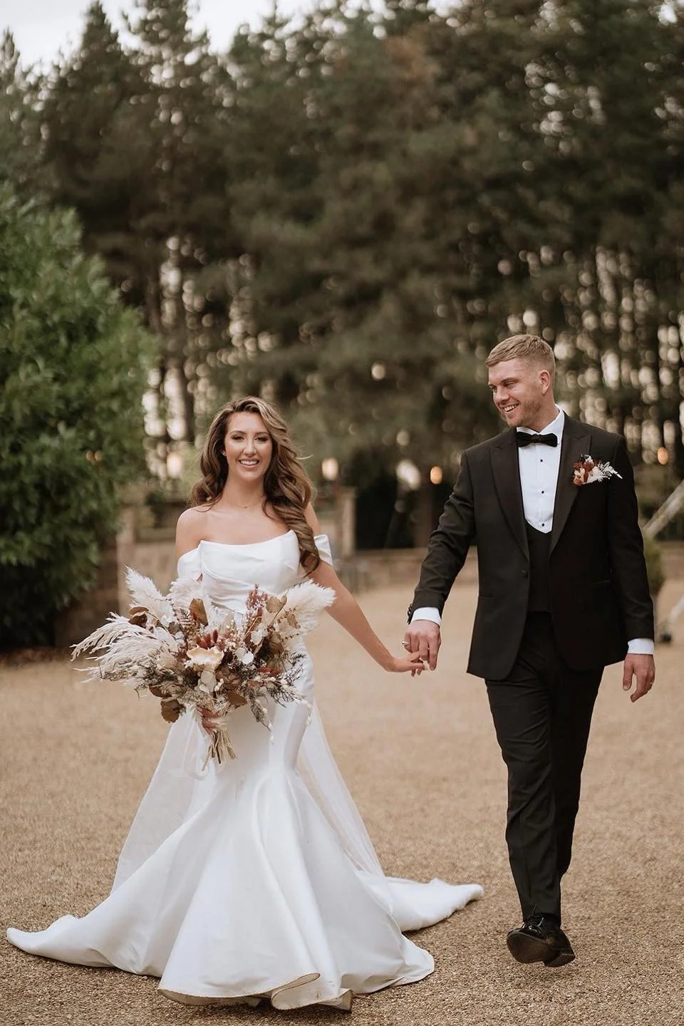 A happy bride and groom walking hand in hand outdoors, smiling, with the bride holding a large bouquet of dried flowers, in a garden or park setting during sunset.