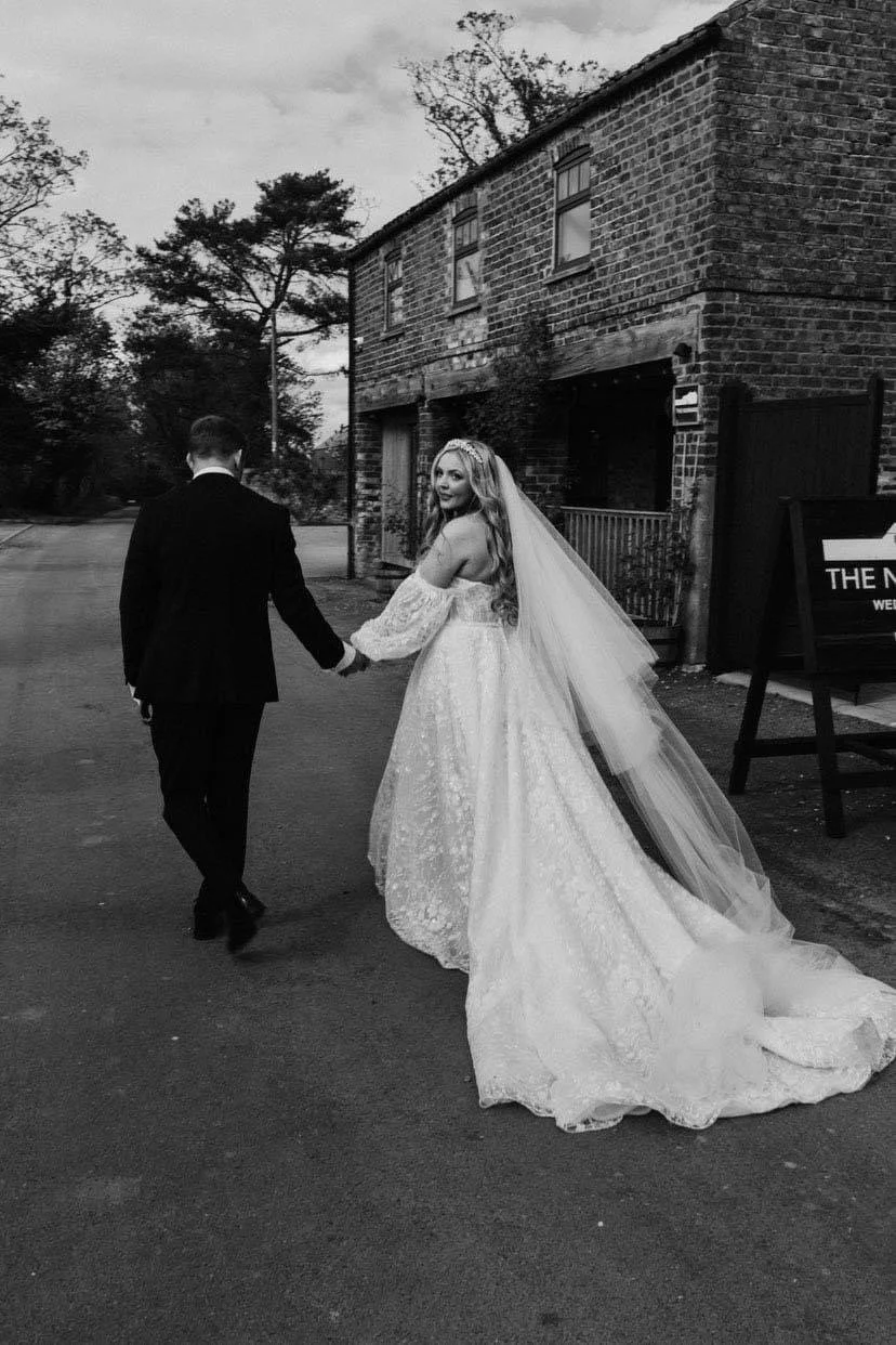A bride and groom holding hands, walking on a street. The bride is in a wedding gown with a long train and veil, and the groom is in a dark suit. They are near a brick building.