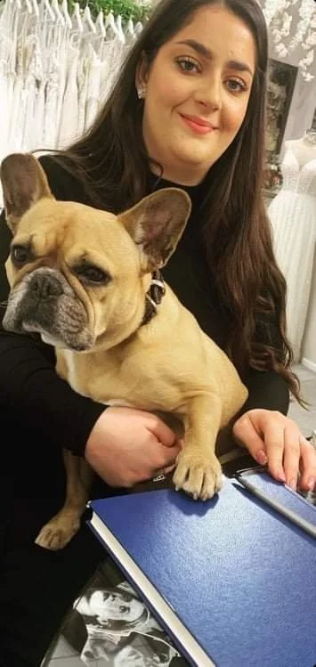 A woman with long dark hair and light skin holding a tan French Bulldog with a wrinkled face in an indoor setting.