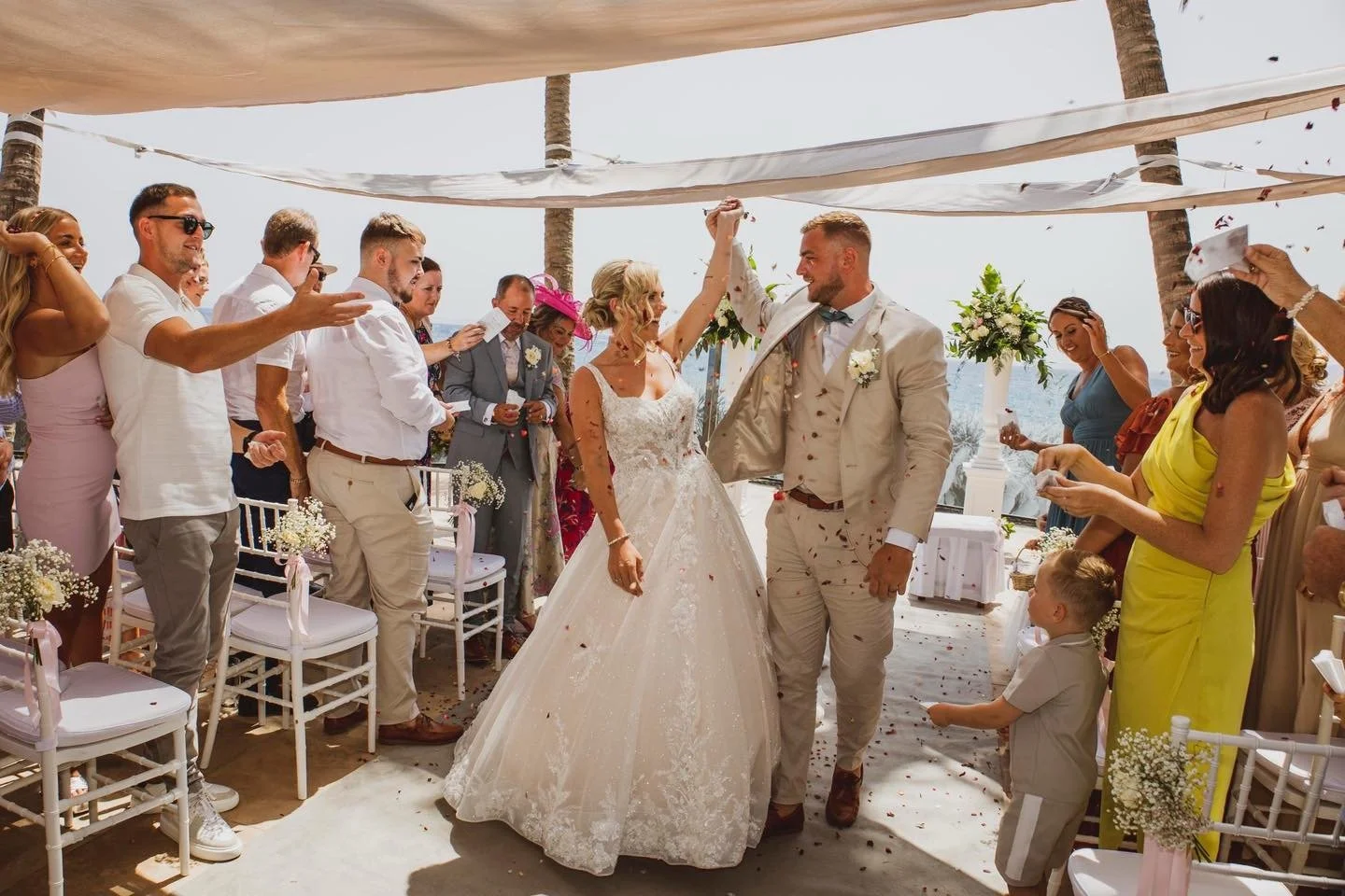 A wedding ceremony takes place outdoors under a canopy with palm trees, overlooking the water. The bride and groom celebrate by holding hands in the center, surrounded by guests. The bride wears a lace wedding gown, and the groom wears a light-colore